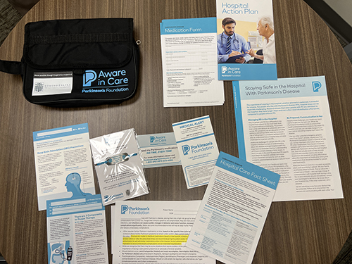 The image shows an "Aware in Care" kit from the Parkinson's Foundation, containing informational pamphlets, forms, and a carrying case designed to help people with Parkinson's disease stay safe in the hospital.
