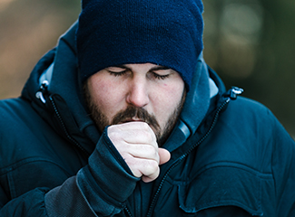 A bearded man wearing a blue jacket and hat is seen coughing.