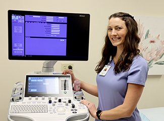 A smiling healthcare professional is standing next to a vascular ultrasound machine.