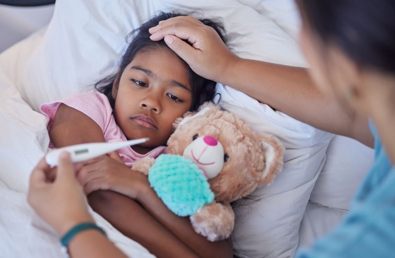 Sick child in bed with stuffed animal while mom checks temperature